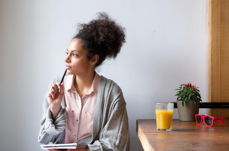 O que levar em conta antes de financiar um imóvel | Foto de uma mulher pensativa, segurando um lápis e um caderno, sentada em uma mesa | Trilha da Conquista | Blog da Tenda