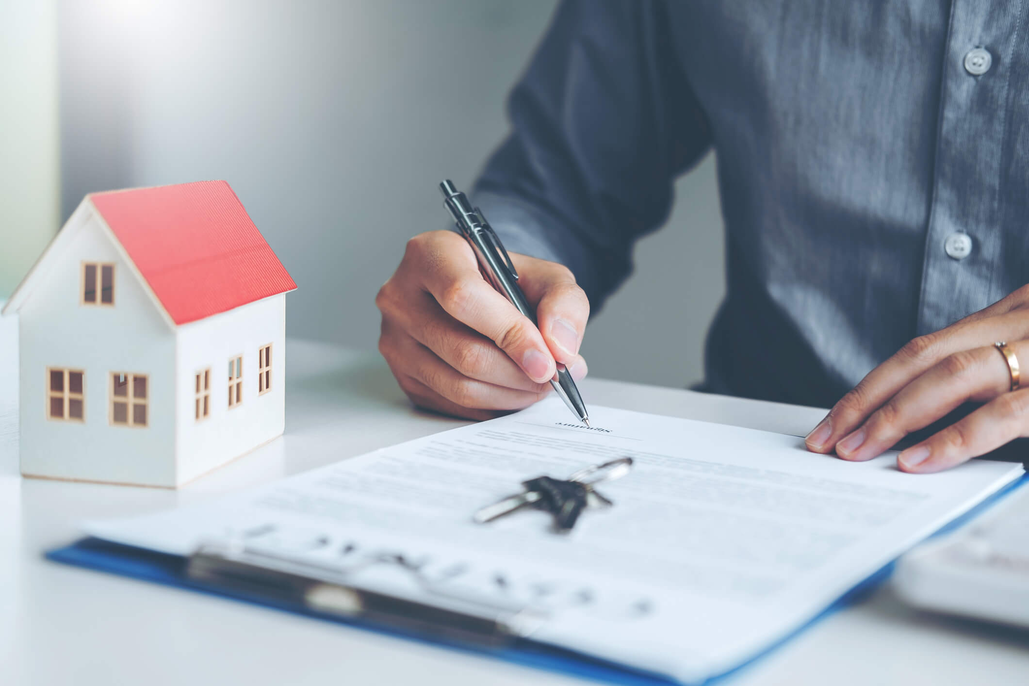 Em uma mesa, homem assina um contrato ao lado de uma casa em miniatura | Diminuir o valor de financiamento imobiliário | Blog da Tenda
