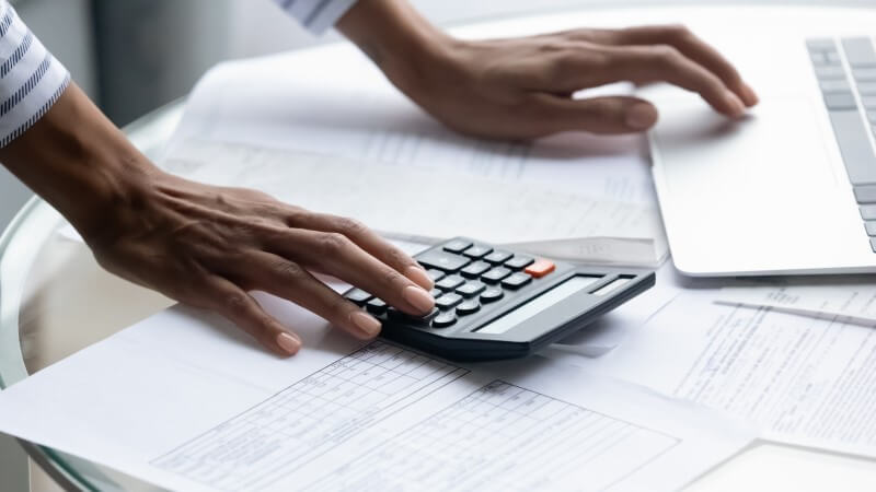 Foto das mãos de uma pessoa fazendo as contas com calculadora e computador | Organização Financeira | Economize | Blog da Tenda