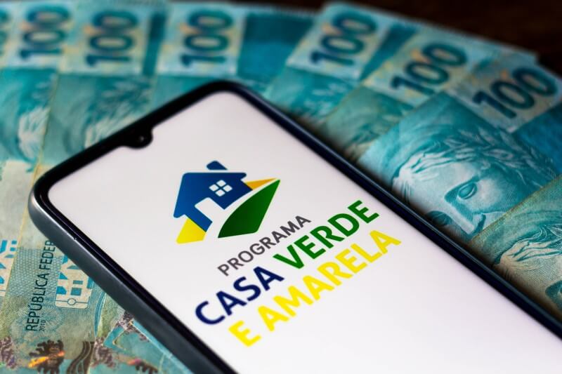 Foto do logo do programa Casa Verde e Amarela em um celular sobre dinheiro | Tudo sobre o programa Casa Verde e Amarela | Guia da Tenda | Blog da Tenda