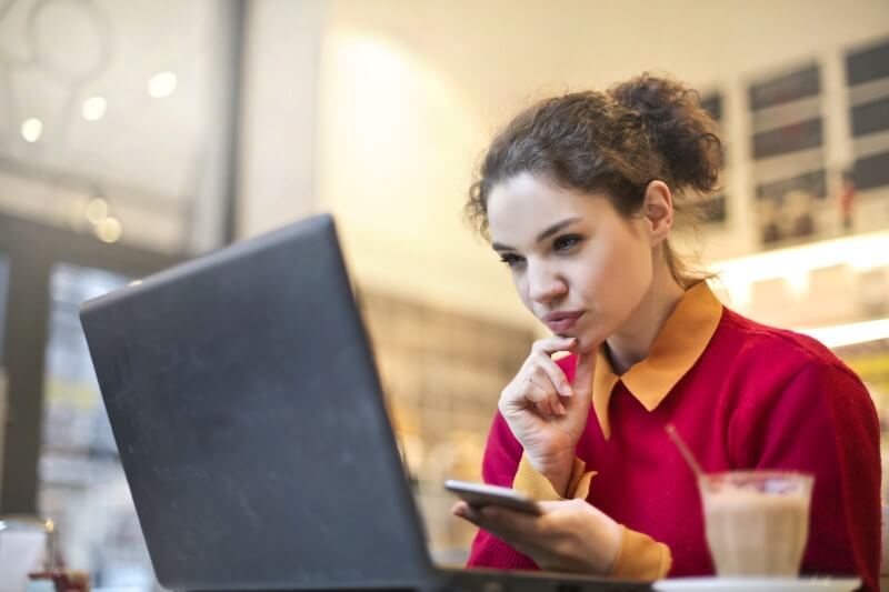 Foto de uma moça com dúvida na frente do computador e com celular na mão | Como comprar um apartamento novo | Guia da Tenda | Blog da Tenda