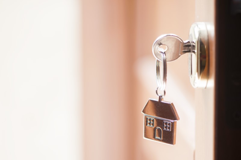 O que é o programa Minha Casa Minha Vida | Foto de um chave com chaveiro de casinha, pendurado em uma porta entreaberta | Minha Casa Minha Vida | Blog da Tenda