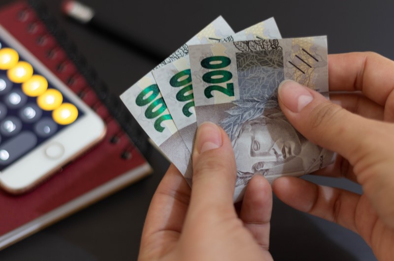 Quais são as vantagens de ter uma renda mista? | Foto de uma pessoa segurando notas de dinheiro com um celular ao fundo | Trilha da Conquista | Blog da Tenda 