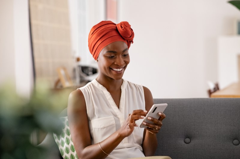 Dicas para fazer uma decoração moderna | Foto de uma mulher sorridente, sentada no sofá enquanto usa o celular | Casa e Decoração | Blog da Tenda 