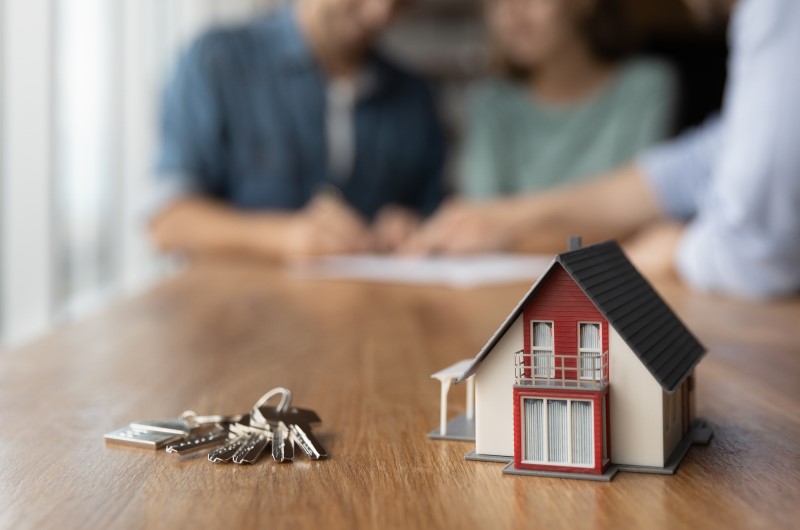 Financiar apartamento na planta | Foto de uma miniatura de casa com chaves do lado e um casal assinando papeis ao fundo | Trilha da Conquista | Blog da Tenda