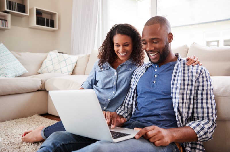 Como dividir ambientes gastando pouco? | Foto de um casal feliz sentados na sala enquanto usam o notebook | Casa e Decoração | Blog da Tenda