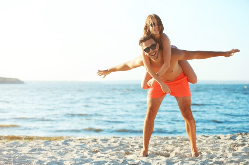 Lugares para visitar em Fortaleza | Foto de uma moça nas costas de um rapaz, sorridentes na areia da praia | Eu Dou Conta | Blog da Tenda 