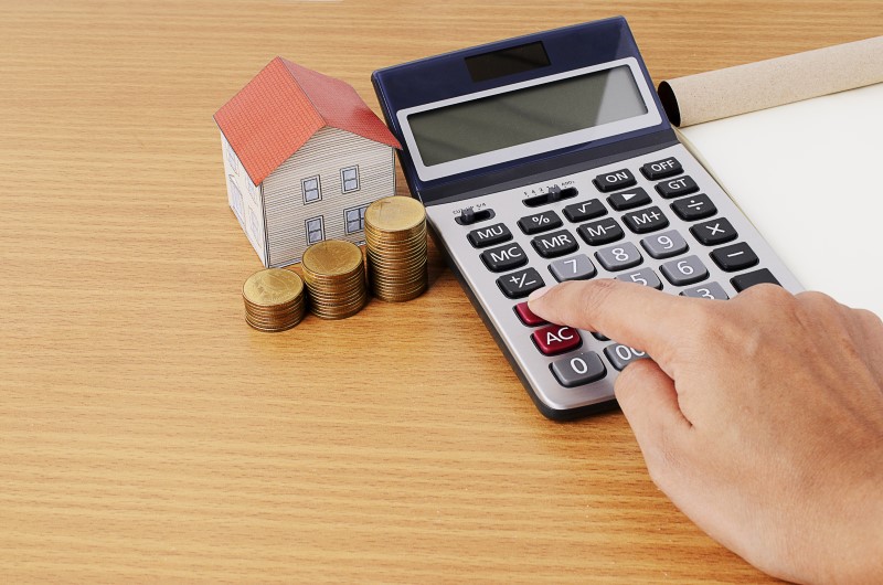 Faixas de renda | Foto de uma pessoa usando calculadora, com moedas e uma casinha ao lado