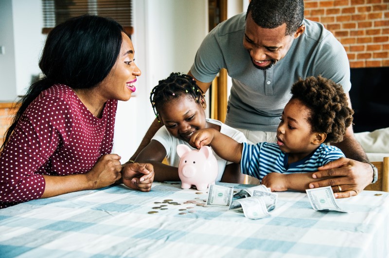 Como organizar o orçamento familiar? | Foto de uma família reunida a mesa, colocando dinheiro em um cofrinho | Economia e Renda Extra | Eu Dou Conta