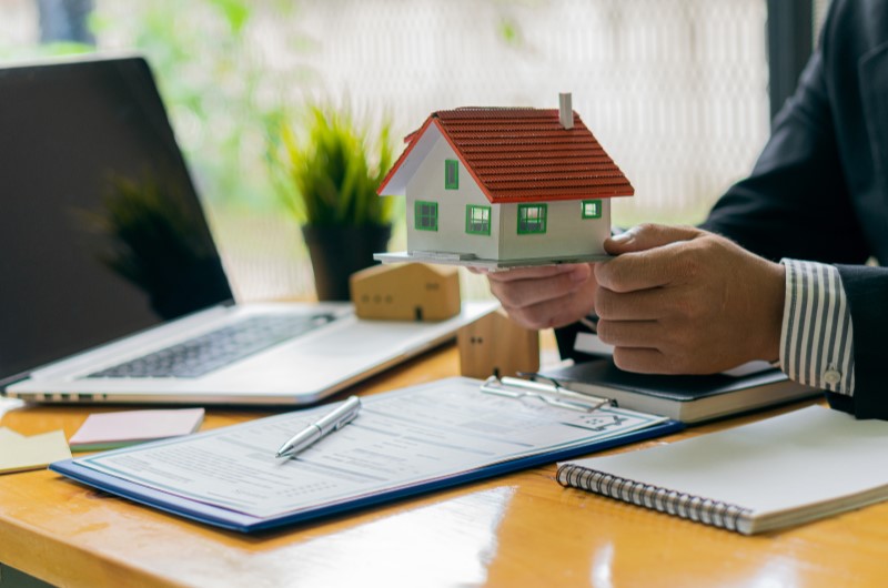 Qual a diferença entre SFH e SFI? | Foto de uma pessoa segurando uma casinha, com um notebook e documentos sobre a mesa | Trilha da Conquista | Blog da Tenda