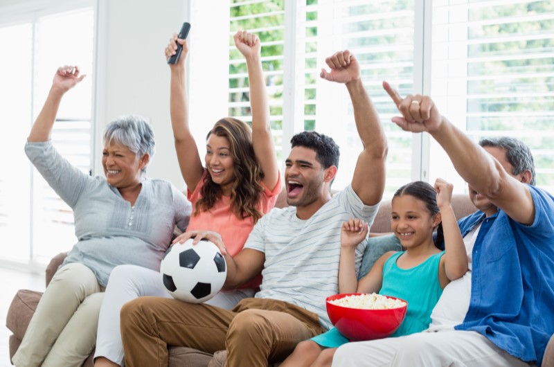 Quando surgiu o futebol? | Foto de uma família no sofá da sala, assistindo futebol | Blog da Tenda