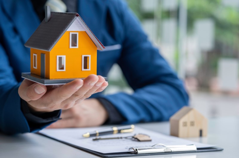 Como comprar um apartamento sem entrada? | Foto de uma pessoa segurando uma casinha em miniatura com alguns documentos sobre a mesa | Trilha da Conquista | Blog da Tenda