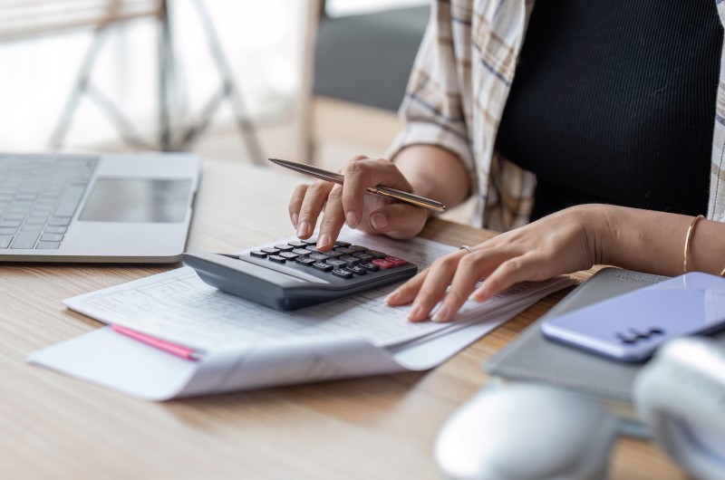 Quais são os tipos de empréstimo? | Foto de uma pessoa usando a calculadora, com alguns papeis e um notebook sobre a mesa | Dúvidas sobre dívidas | Eu Dou Conta