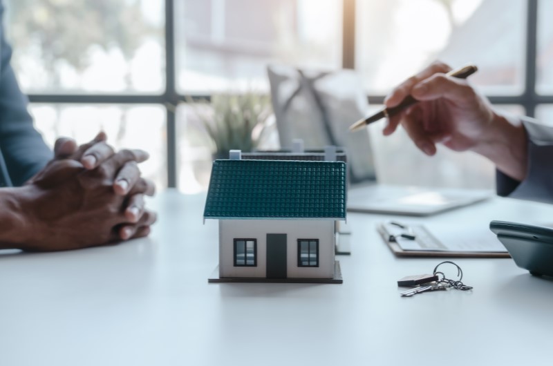 Quando optar pela compra conjunta de imóvel? | Foto de duas pessoas conversando ao fundo e uma casinha em miniatura na frente | Trilha da Conquista | Blog da Tenda