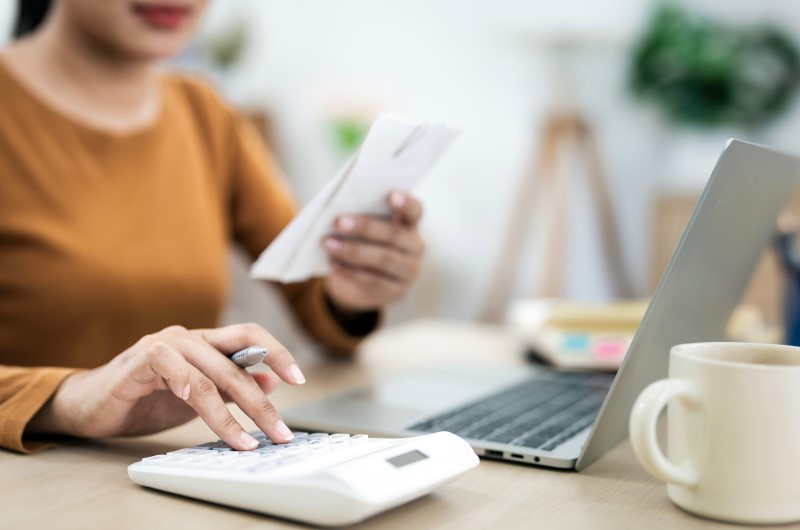 Vida financeira instável | Foto de uma mulher segurando um papel enquanto digita na calculadora | Trilha da Conquista | Blog da Tenda