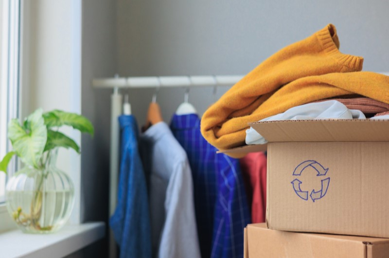 O que é consumo consciente? | Foto de uma caixa com um símbolo de reciclável e roupas ao fundo | Eu Dou Conta | Blog da Tenda