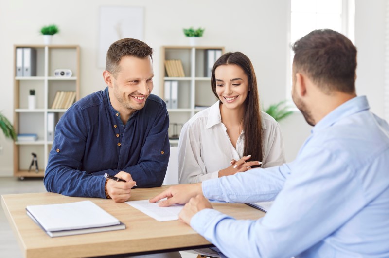 Como sair do aluguel e conquistar a casa própria | Casal sorrindo enquanto revisa documentos com um consultor imobiliário | Trilha da Conquista | Blog da Tenda