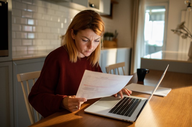 Existe multa rescisória | Mulher analisando documentos em frente a um notebook em uma mesa de cozinha | Trilha da Conquista | Blog da Tenda 