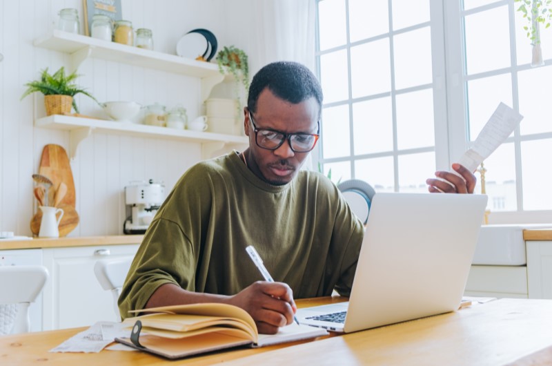 Anote todas as receitas e despesas | Homem segurando recibos, anotando em caderno e usando notebook | Eu Dou Conta | Blog da Tenda 