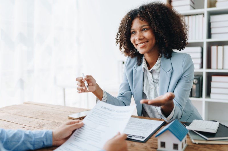 Análise de crédito | Corretora explicando contrato para cliente com miniatura de casa na mesa | Trilha da Conquista | Blog da Tenda 