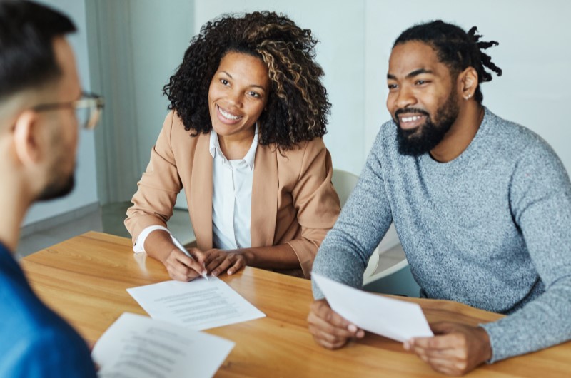 Diferença entre consórcio e financiamento imobiliário | Casal sorridente conversando com um corretor de imóveis | Trilha da Conquista | Blog da Tenda