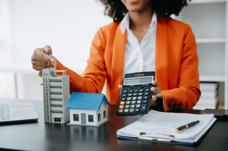 O que são despesas ordinárias e extraordinárias de condomínio | Mulher com calculadora e maquetes de imóveis sobre a mesa | Viver em Condomínio | Blog da Tenda 