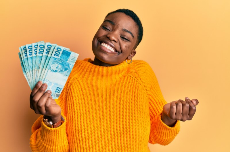 O que é renda extra | Mulher sorridente com notas de 100 reais na mão | Economia e renda extra | Eu Dou Conta