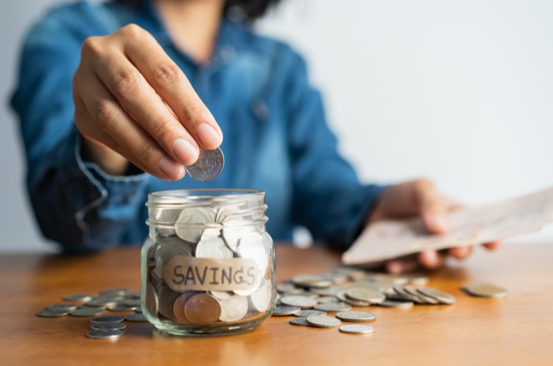 Como cortar custos | Foto de uma mulher colocando moedas em um pote | Economia e renda extra | Eu Dou Conta