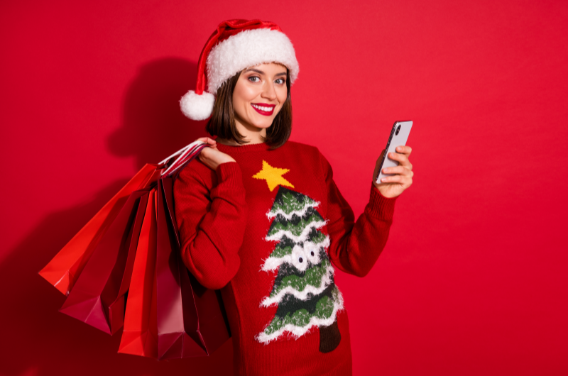 Como economizar nos presentes de Natal | Foto de uma mulher com gorro de papai noel segurando sacolas de compras e um celular | Economia e renda extra | Eu Dou Conta
