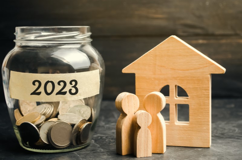 Organização financeira | Foto de um cofrinho com moedas para 2023, uma casa de madeira e uma família de madeira | Como limpar o seu nome | Eu Dou Conta
