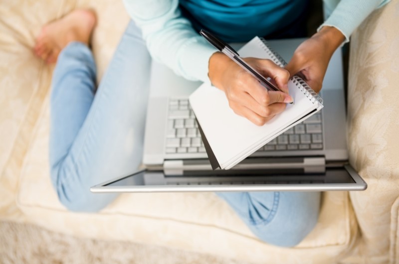 Faça um planejamento financeiro | Foto de uma pessoa sentada com um notebook no colo, enquanto faz anotações em um papel | Economia e renda extra | Blog Eu Dou Conta 