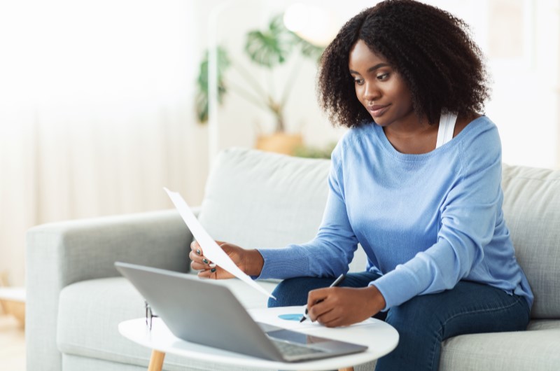 Como é feita a análise de crédito? Foto de uma mulher sentada no sofá e segurando um papel enquanto usa o notebook | Dúvidas sobre dívidas | Eu Dou Conta
