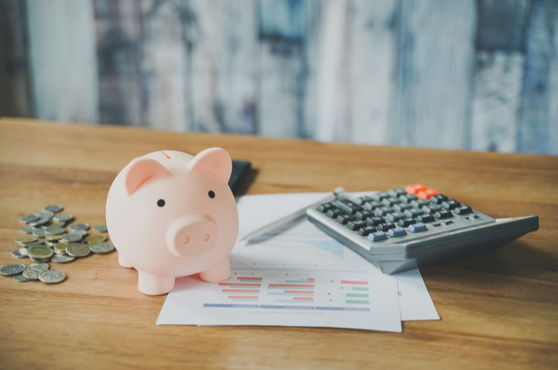 Como investir com pouco dinheiro? | Foto de um cofrinho em cima de uma papel, ao lado de moedas e calculadora | Economia e renda extra | Eu Dou Conta 