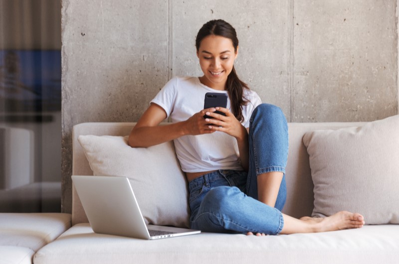 Mantenha os seus dados atualizados | Foto de uma mulher sentada no sofá e olhando para o celular, com o notebook ao lado | Dúvidas sobre dívidas | Eu Dou Conta 