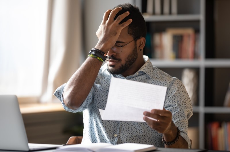 O que fazer se minha análise de crédito for reprovada? | Fotot de um homem na frente de um notebook, enquanto segura um papel e coloca a mão no rosto | Dúvidas sobre dívidas | Eu Dou Conta