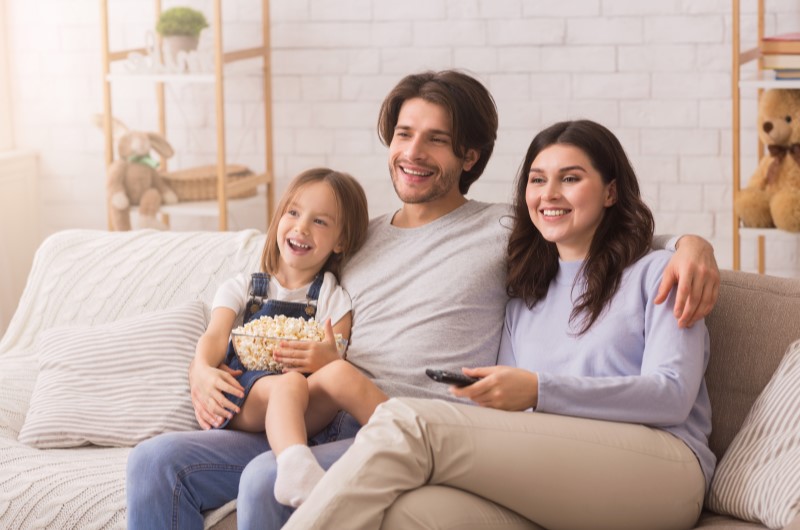 Cuidado com televisões e aparelhos em stand by | Foto de uma família feliz sentada no sofá e assistindo televisão | Economia e renda extra | Eu Dou Conta 