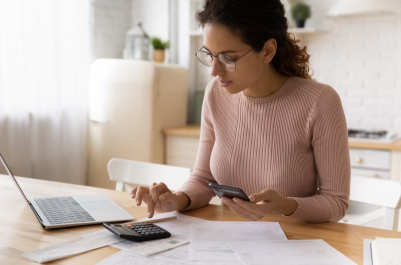Cuidados para economizar energia em casa | Foto de uma mulher usando uma calculadora, com um notebook e alguns papeis ao redor | Economia e renda extra | Eu Dou Conta 