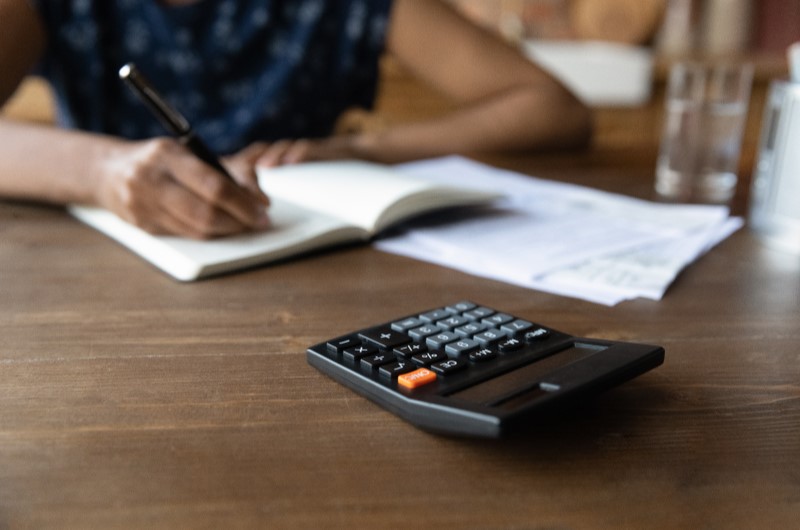 Use o método 50-30-20 | Foto de uma pessoa anotando em um caderno, com uma calculadora ao lado | Como limpar o seu nome | Eu Dou Conta