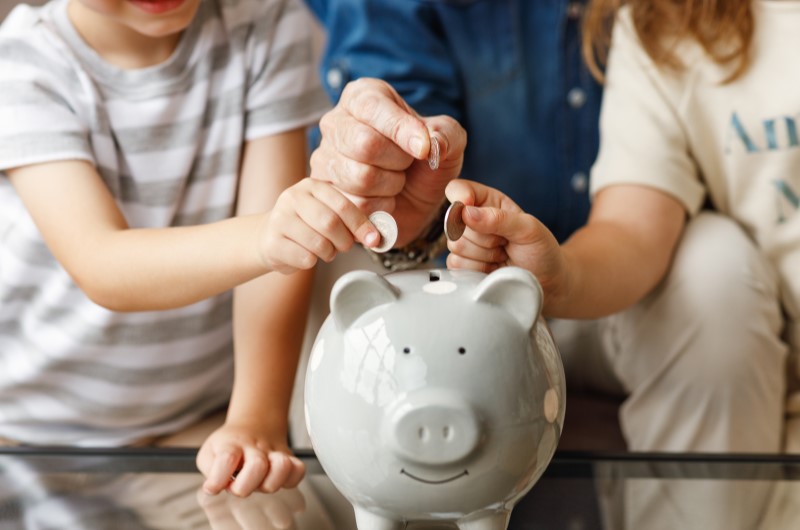 Como ter uma boa educação financeira? | Foto de um adulto e duas crianças colocando moedas em um cofrinho | Economia e renda extra | Eu Dou Conta 