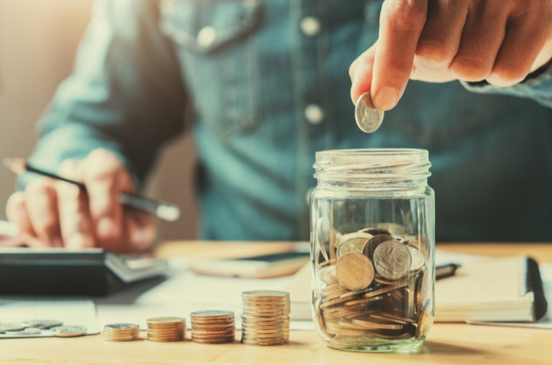 Invista seu dinheiro | Foto de uma pessoa usando uma calculadora e colocando moedas em um pote | Economia e renda extra | Eu Dou Conta 