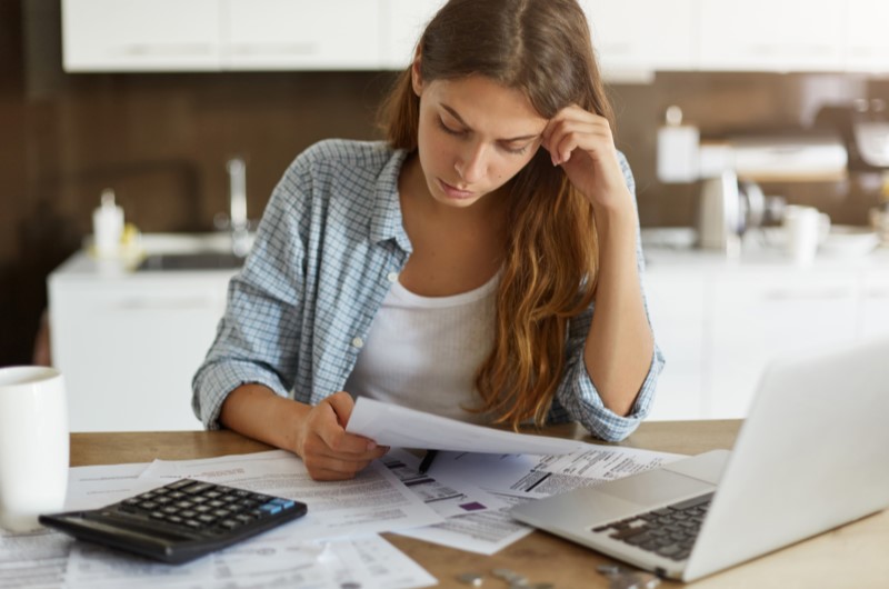 3 dicas para organizar esses gastos | Foto de uma mulher concentrada olhando para um papel, com uma calculadora e notebook sobre a mesa | Economia e renda extra | Eu Dou Conta 