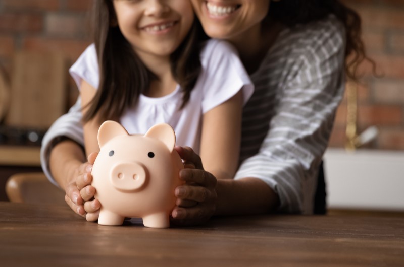 O que é educação financeira? | Foto de uma mãe e filha sorridentes segurando um cofrinho | Economia e renda extra | Eu Dou Conta 