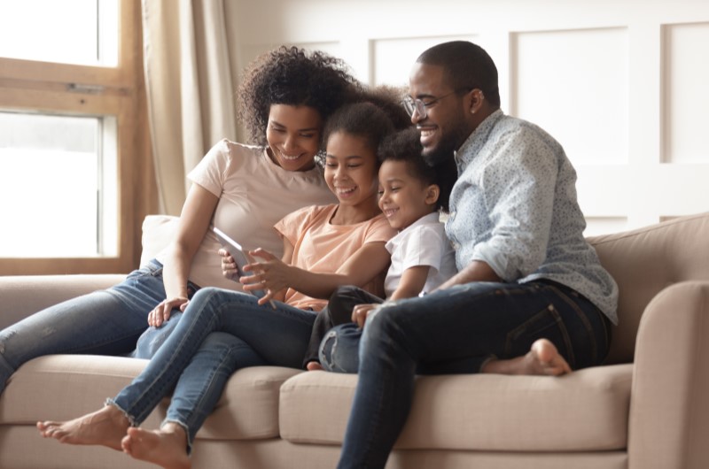 Pesquise por vídeos e desenhos infantis sobre o tema | Foto de uma família feliz assistindo um vídeo na sala de estar | Economia e renda extra | Eu Dou Conta 