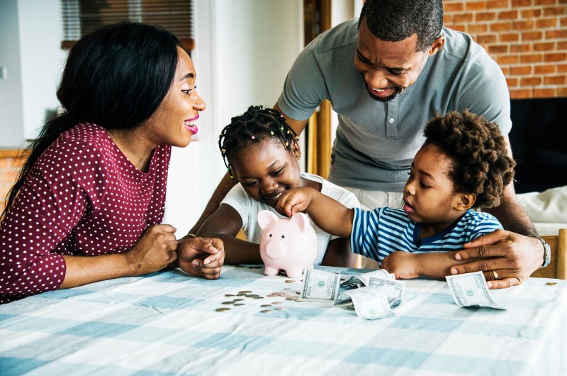 Como organizar o orçamento familiar? | Foto de uma família reunida a mesa, colocando dinheiro em um cofrinho | Economia e Renda Extra | Eu Dou Conta 