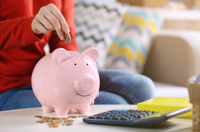 Como se organizar financeiramente: faça uma renda extra | Foto de uma pessoa colocando moedas em um cofrinho, com uma calculadora ao lado | Dúvidas sobre dívidas | Eu Dou Conta 