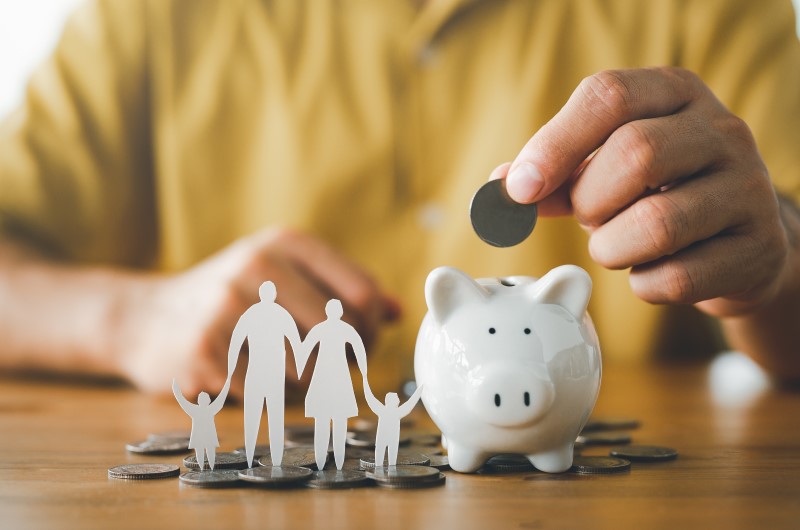 O que é um orçamento familiar? | Foto de uma pessoa colocando uma moeda no cofrinho, com moedas e uma família de papel ao lado | Economia e Renda Extra | Eu Dou Conta 