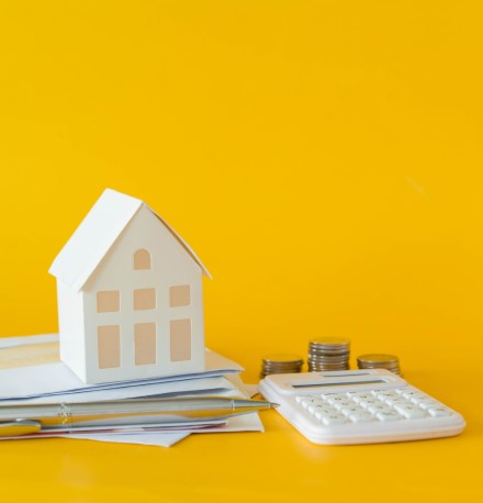 Amortização de financiamento imobiliário | Foto de um fundo amarelo, com uma casinha, alguns papeis e moedas, e uma calculadora | Dúvidas sobre dívidas | Eu Dou Conta
