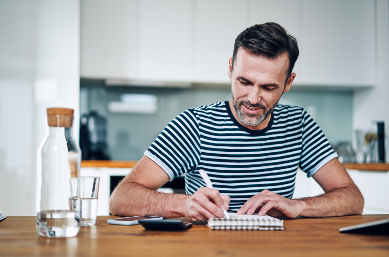 Diferenças entre Tabela SAC e Tabela Price | Foto de um homem escrevendo em um caderno, com uma calculadora ao lado | Dúvidas sobre dívidas | Eu Dou Conta 