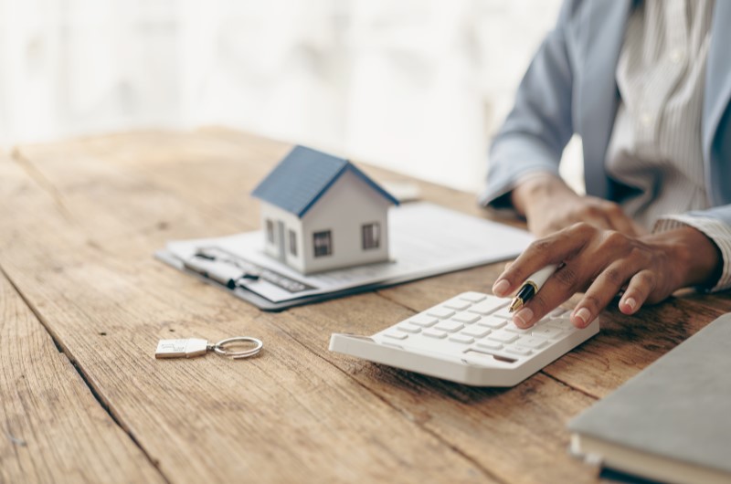 É preciso declarar imóvel financiado? | Foto de uma pessoa usando a calculadora com um casinha e documento ao lado | Dúvidas sobre dívidas | Eu Dou Conta 